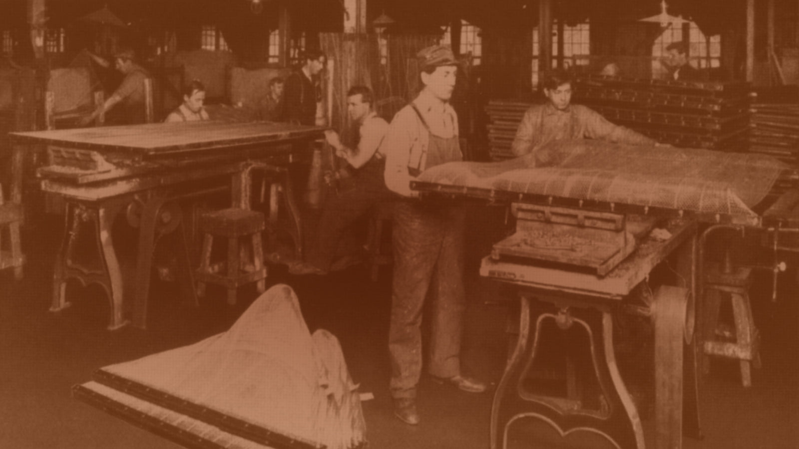 Sepia-toned early-1900s photograph of young factory workers hand-building mattresses at long wooden worktables.